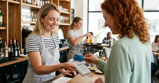 Eine lächelnde Barista nimmt an der Theke eines Cafés eine Kreditkartenzahlung von einer Kundin entgegen.