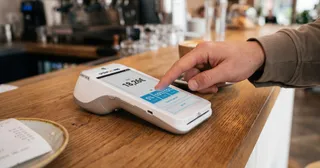 Eine Person bedient ein Zahlungsterminal mit Touchscreen auf einer Theke und schließt gerade eine Transaktion über 18,24 € plus Trinkgeld ab.