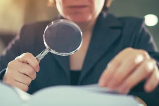 Eine Finanzprüferin hält eine Lupe vor Papiere nd prüft die Dokumente aufmerksam.